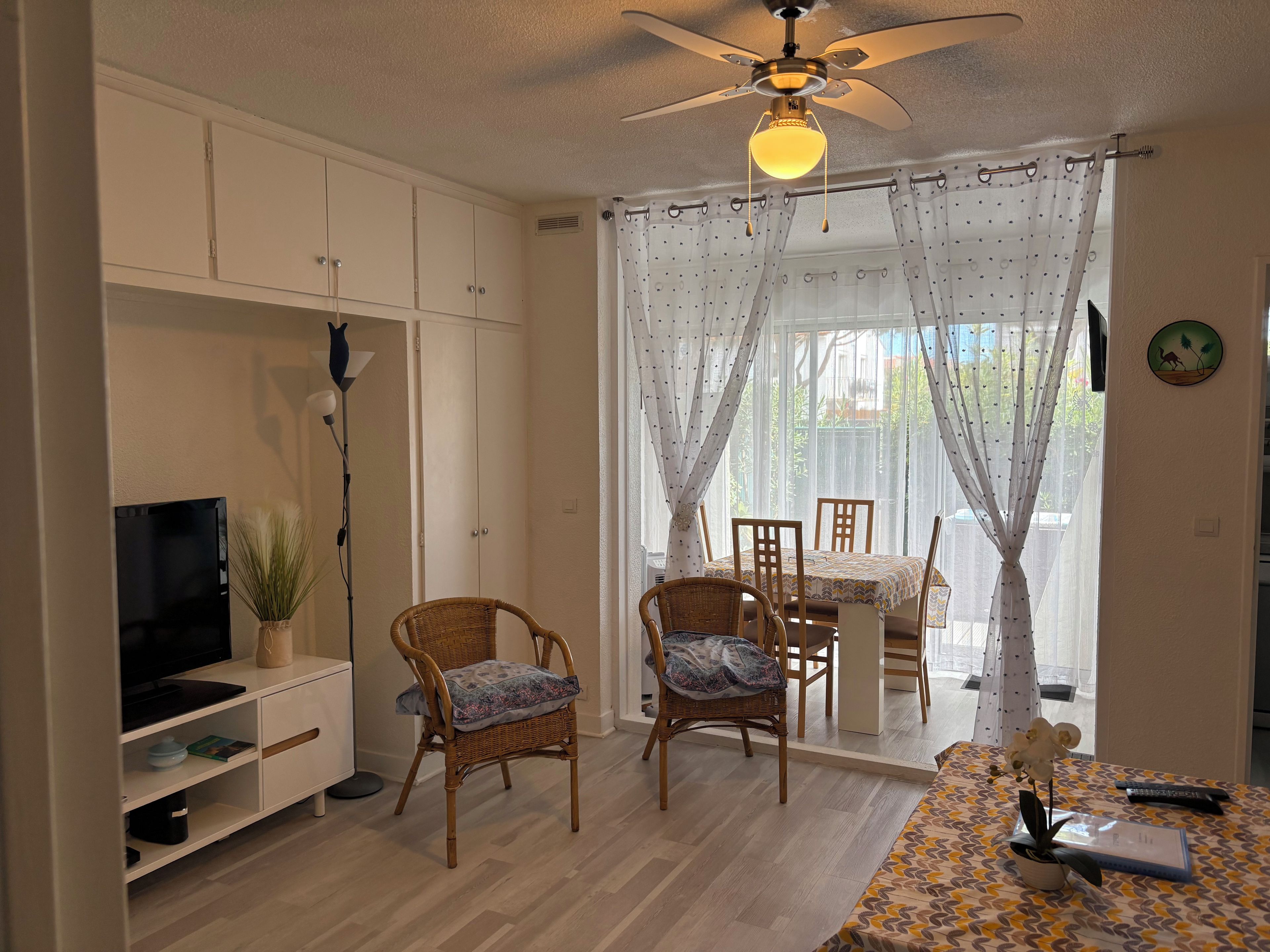 A room with a ceiling fan, a television on a stand, wicker chairs, a dining table with chairs, and a window with curtains.