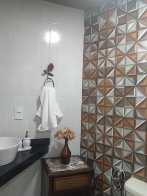Casa de banho com lavatório branco, um porta-toalhas com toalha branca, um vaso com flores, uma mesinha de cabeceira de madeira e uma sanita com chuveiro.