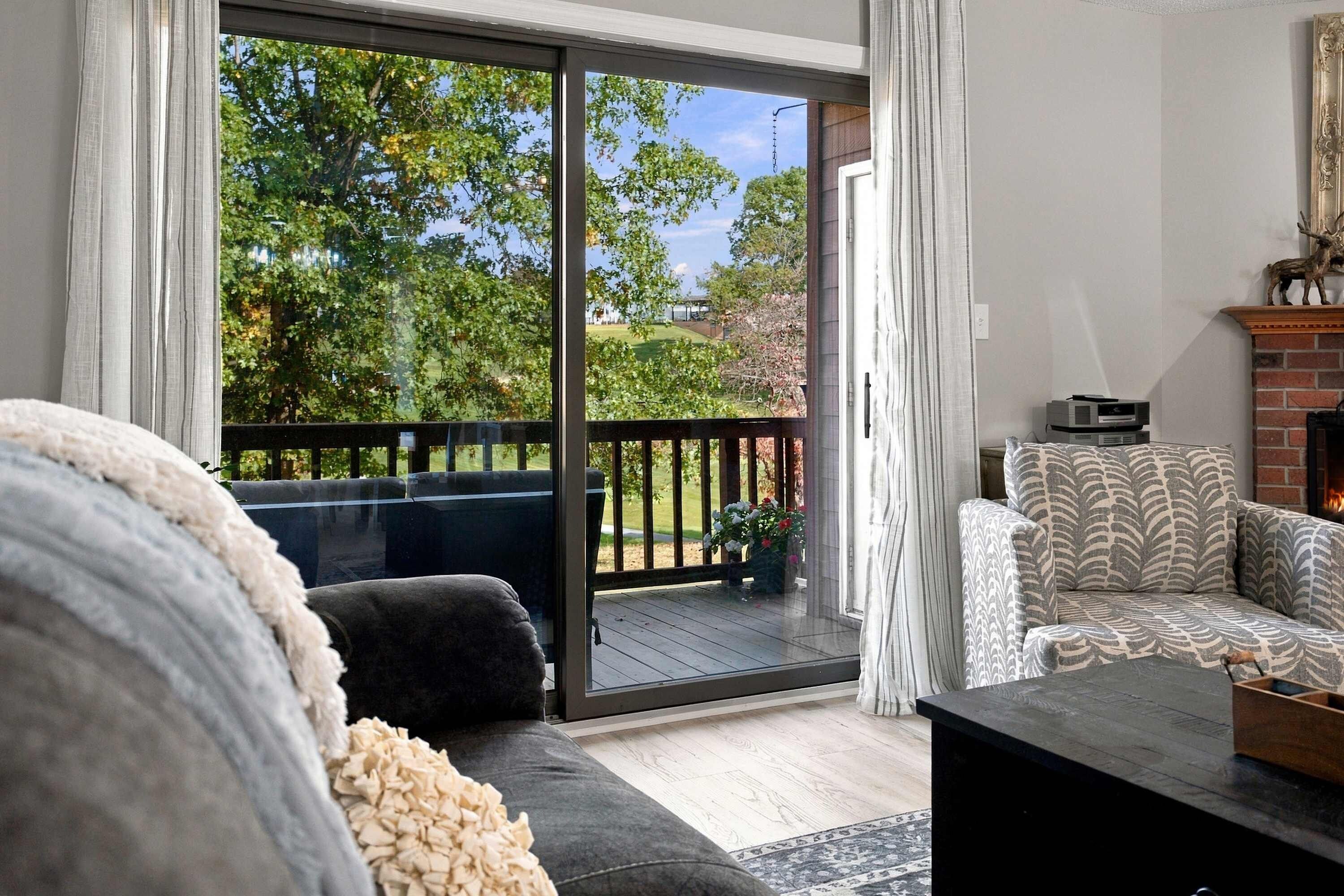 A living room with a sofa, a patterned armchair, a fireplace, and a sliding glass door leading to a deck with a view of trees and a lawn.