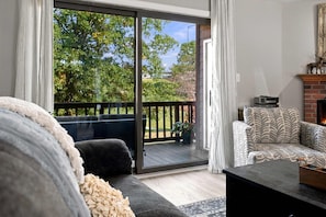 A living room with a sofa, a patterned armchair, a fireplace, and a sliding glass door leading to a deck with a view of trees and a lawn.