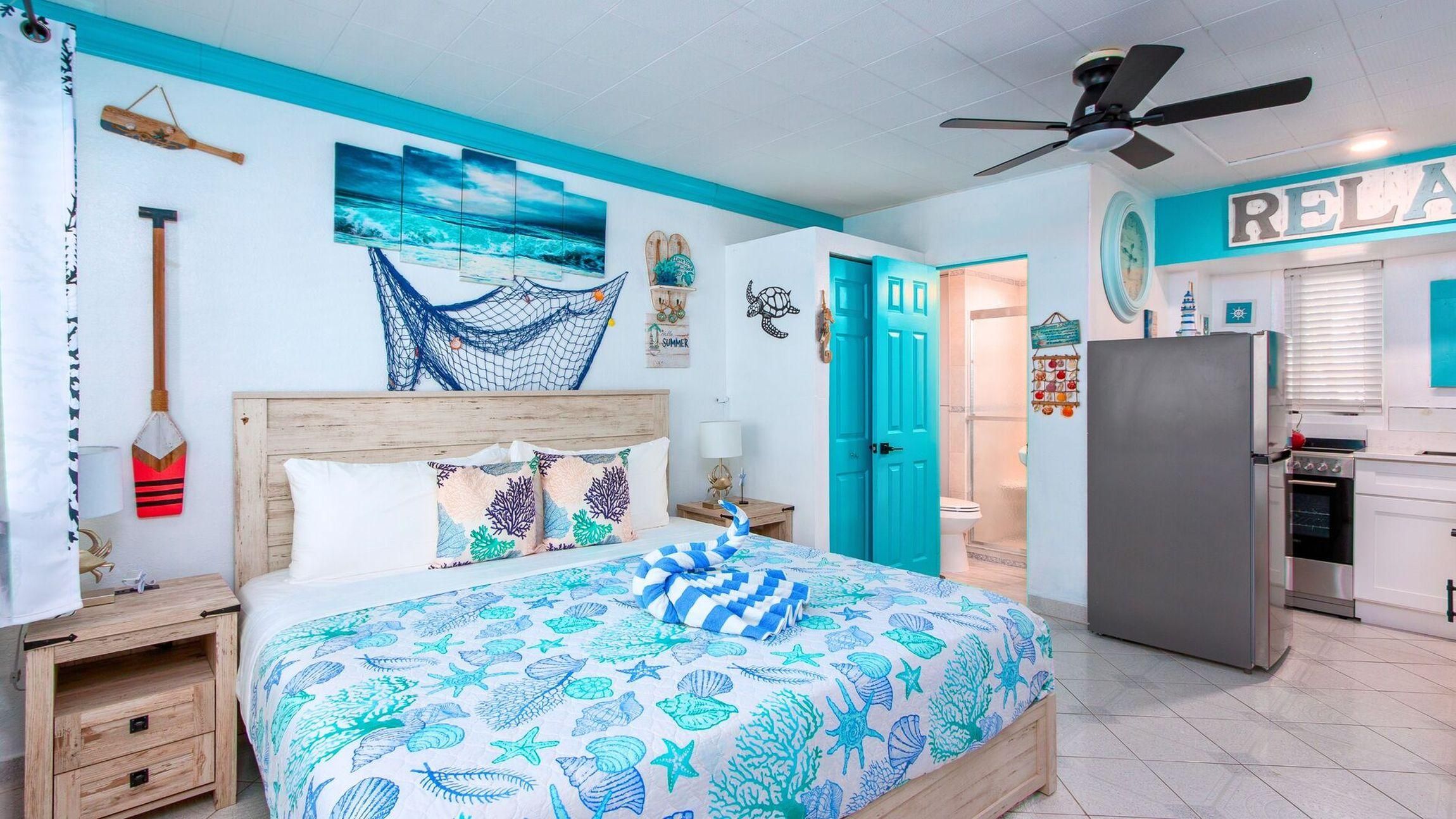 Un dormitorio con cama, mesita de noche, cabecera de madera, ventilador de techo y una zona de cocina con refrigerador.