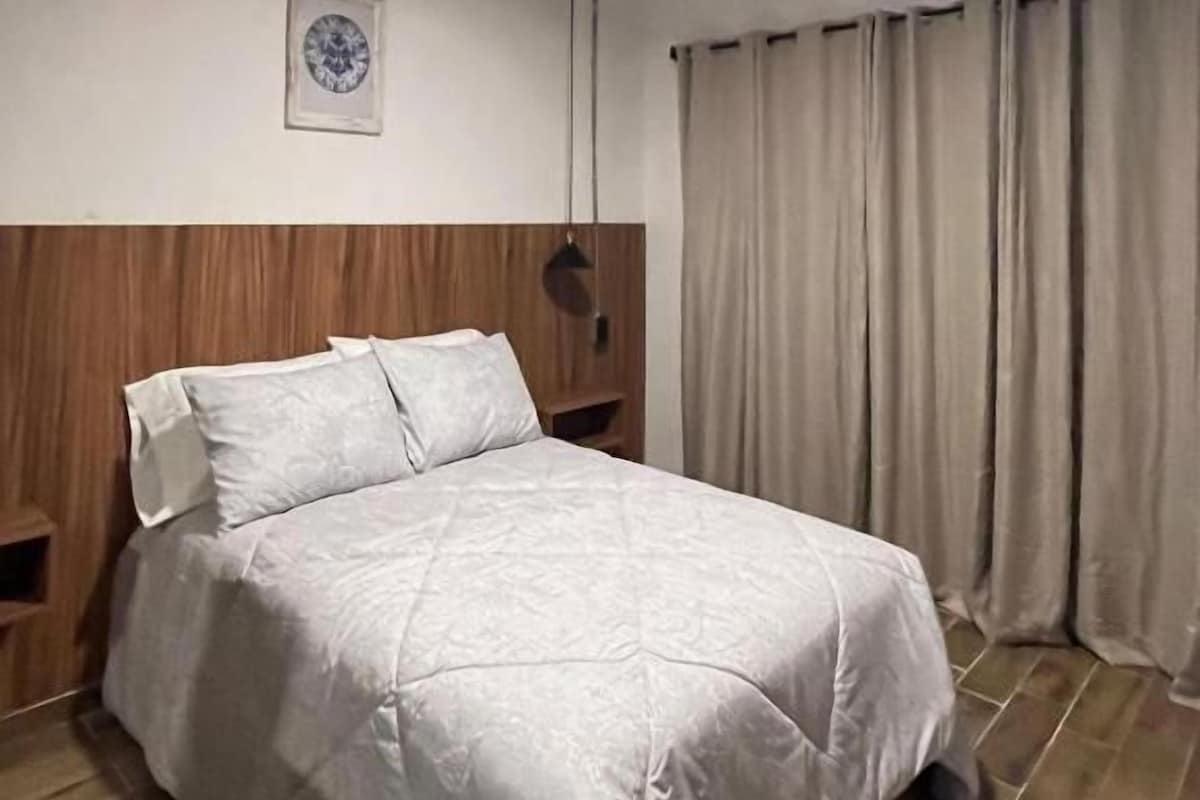 Quarto de hotel com cama, cabeceira de madeira, luminária fixada na parede, quadro emoldurado e janela com cortinas.