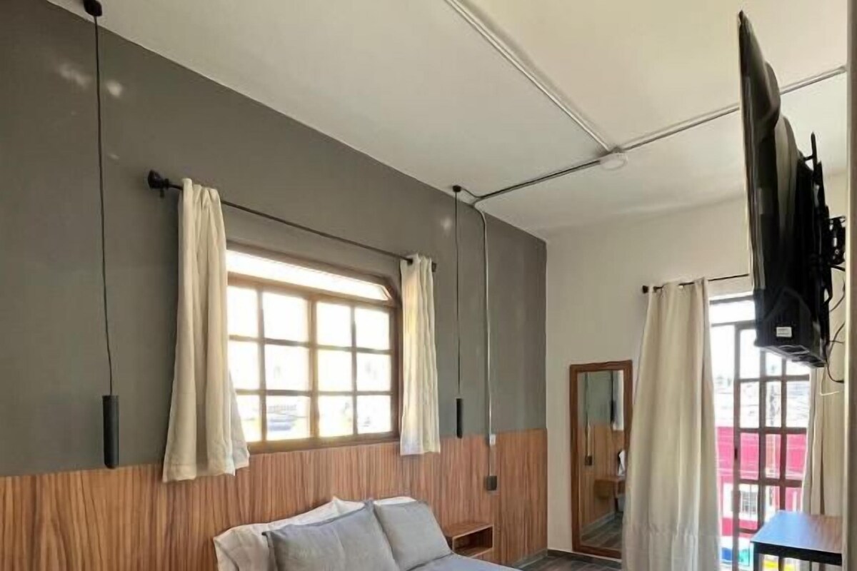 Quarto com cama, painéis de madeira, janela com cortinas, televisão e porta.