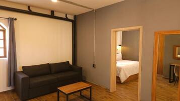A room with a dark-colored sofa, a small wooden table, a bed with white bedding, and a mirror on the wall.