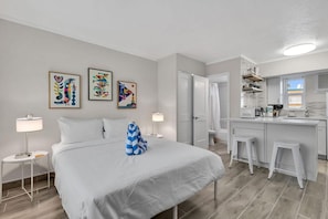 Ett modernt hotellrum med en säng, sänglampor, ett skrivbord med en stol, ett badrum och ett litet kök.
