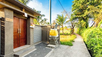 A villa entrance with a wooden door, a signboard, a small garden area, and a pathway leading to the villa.