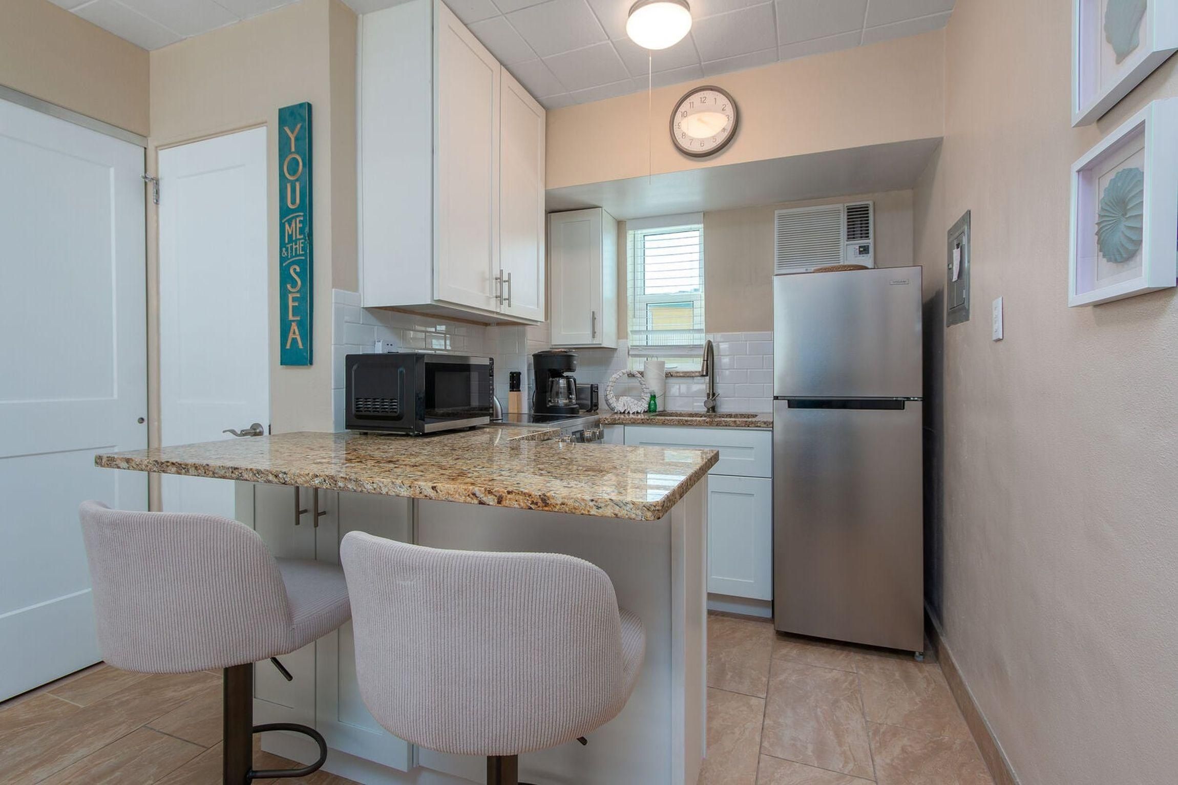 A compact kitchen with a microwave, coffee maker, and refrigerator. There are two chairs and a clock on the wall.