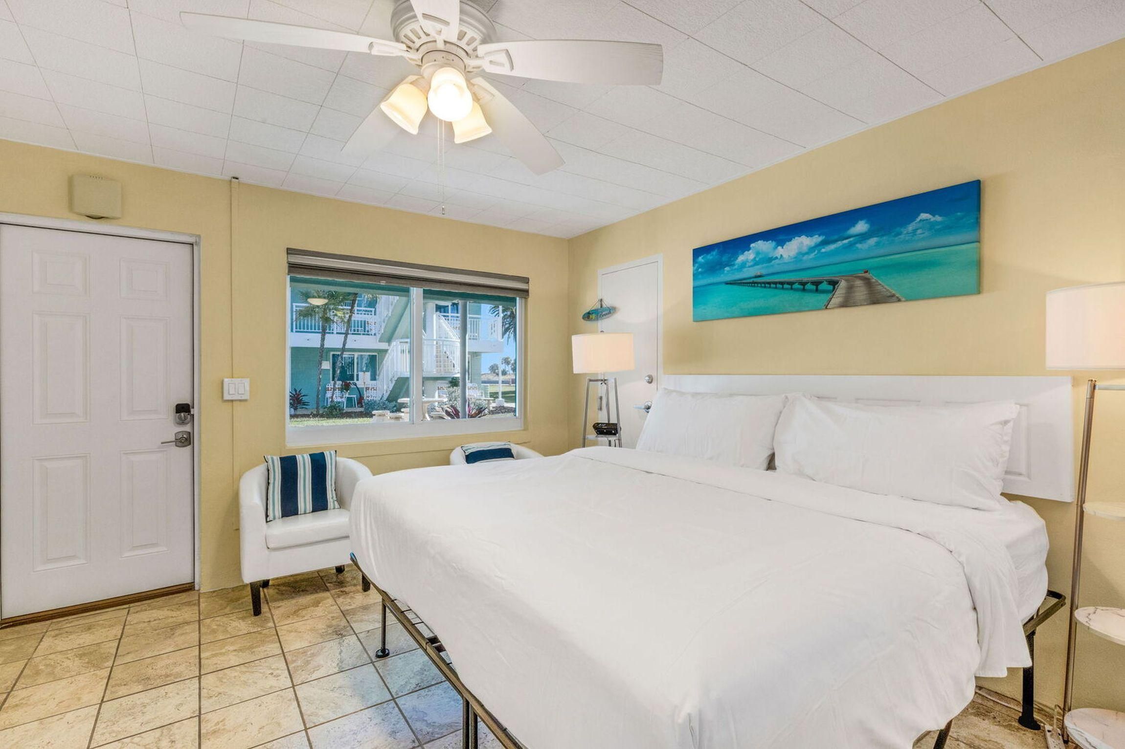Habitación de hotel con cama, ventana con vista a un edificio, ventilador de techo y una pintura de un muelle.