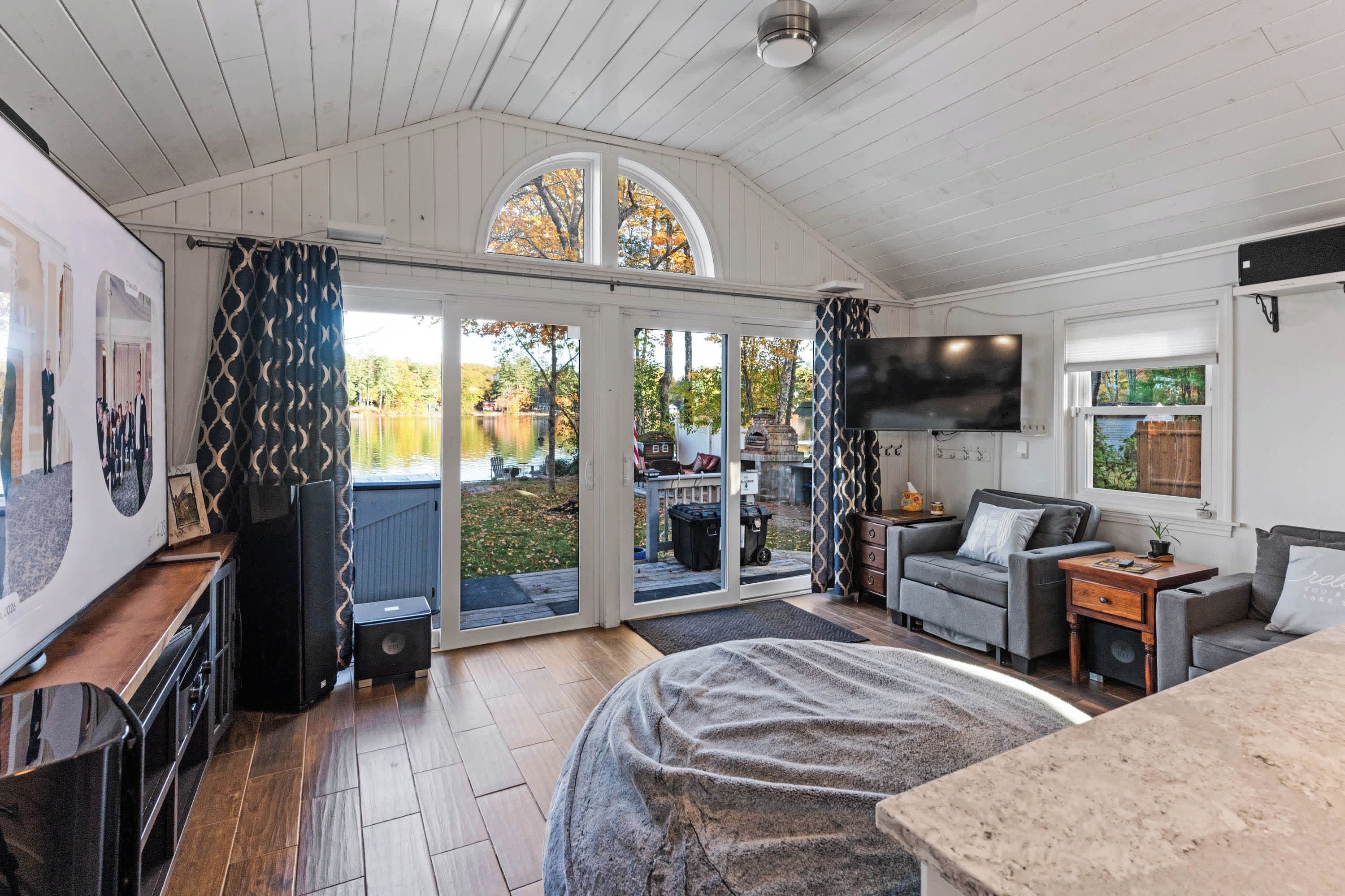 A cozy room with a large bed, a television, a sofa, and a sliding glass door leading to an outdoor area with a view of trees and a body of water.