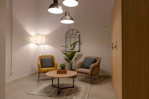 Una habitación con una mesa redonda, dos sillas, una lámpara de pie, una planta en maceta y un espejo montado en la pared.