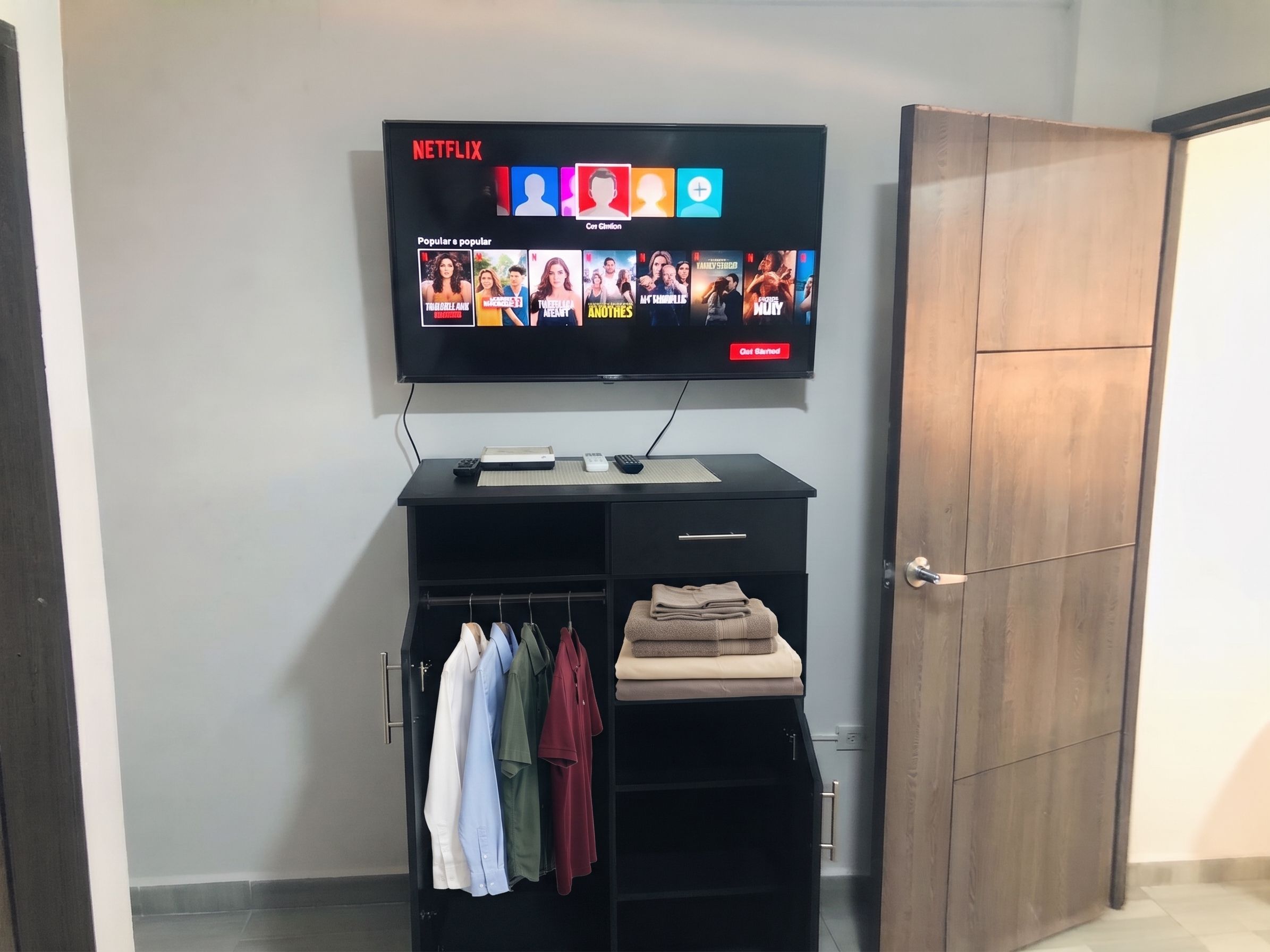 A flat-screen TV mounted on a wall displaying various Netflix shows. Below the TV, there is a black entertainment unit with a remote control.