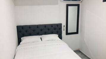 Une chambre d’hôtel étroite, avec un lit simple, une tête de lit capitonnée de couleur sombre, un climatiseur fixé au mur et un grand miroir.