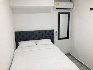 Kapea hotellihuone, jossa on yksi sänky, tumma, kuviollinen pääty, seinään asennettu ilmastointilaite ja suuri peili.
