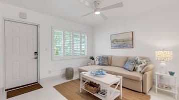 Ein geräumiges Wohnzimmer mit einer weißen Tür, einem Deckenventilator, einem beige farbenen Sofa mit gemusterten Kissen, einem Couchtisch und einem gerahmten Bild an der Wand.
