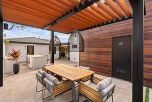 Un espace repas extérieur moderne, avec une table et des chaises en bois, une plante en pot et, au fond, un coin cuisine.