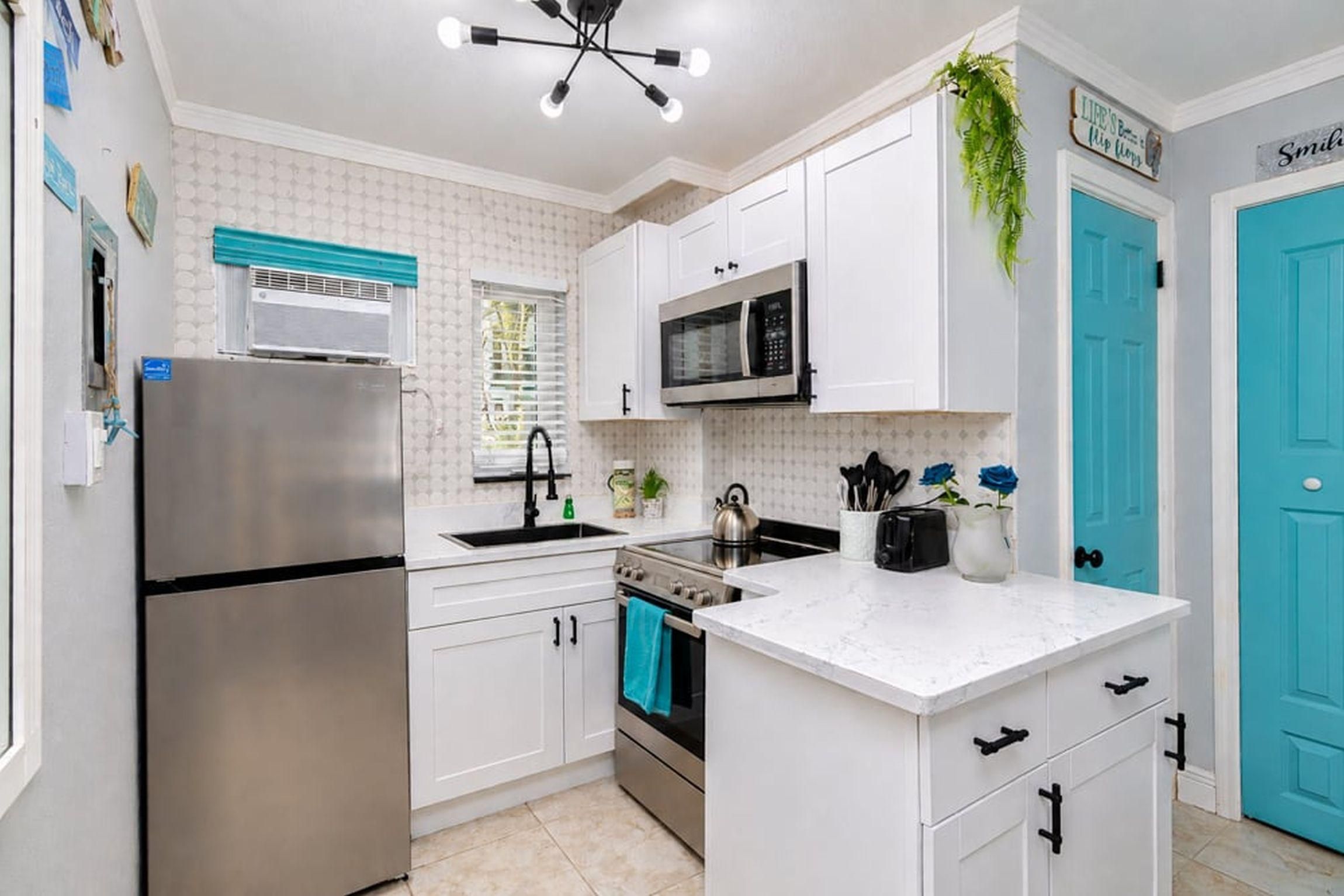 A kitchen with white cabinets, a stainless steel refrigerator, a microwave, and a stove. There is a window with blinds, a ceiling light fixture, and a blue door.