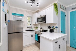 Una cocina con armarios blancos, un refrigerador de acero inoxidable, un microondas y una estufa. Hay una ventana con persianas, un lámpara en el techo y una puerta azul.