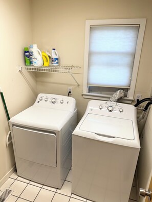 Un cuarto de lavandería con dos lavadoras, un estante con productos de limpieza y una ventana con persianas.