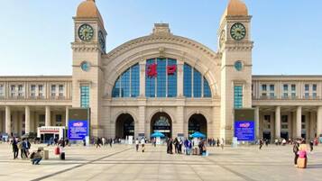 Een groot treinstation met een centrale boogvormige ingang, twee klokkentorens en een ruim plein met mensen en paraplu's.
