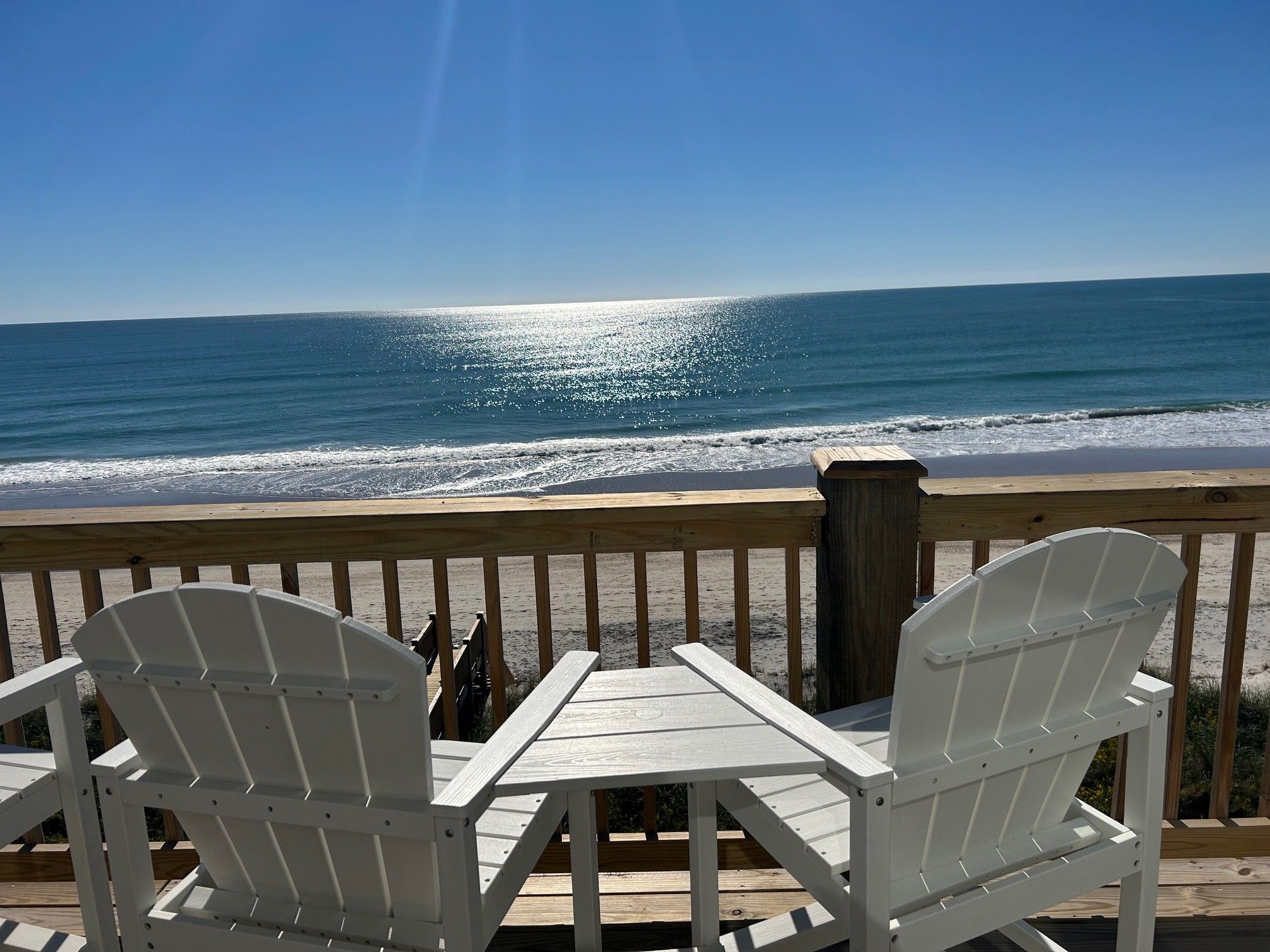 A wooden deck with two white Adirondack chairs facing a beach.