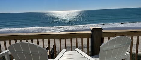 A wooden deck with two white Adirondack chairs facing a beach.