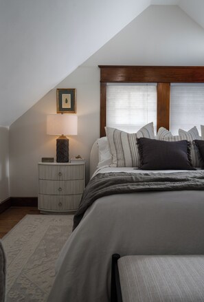 Un dormitorio con techo inclinado, una cama con varias almohadas, una mesita de noche con una lámpara y un cuadro enmarcado en la pared.