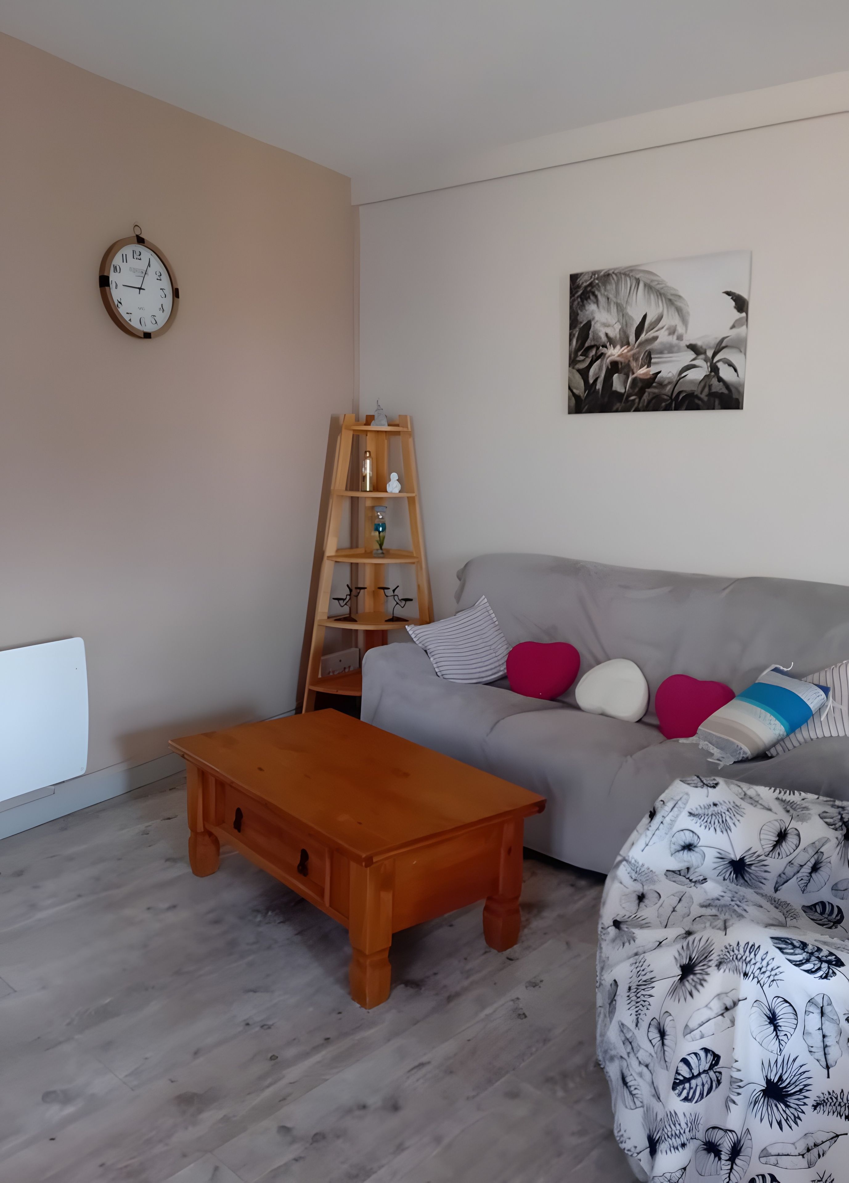A living room with a wooden coffee table, a grey sofa with colorful cushions, a wooden ladder shelf, a wall clock, and a floral artwork.