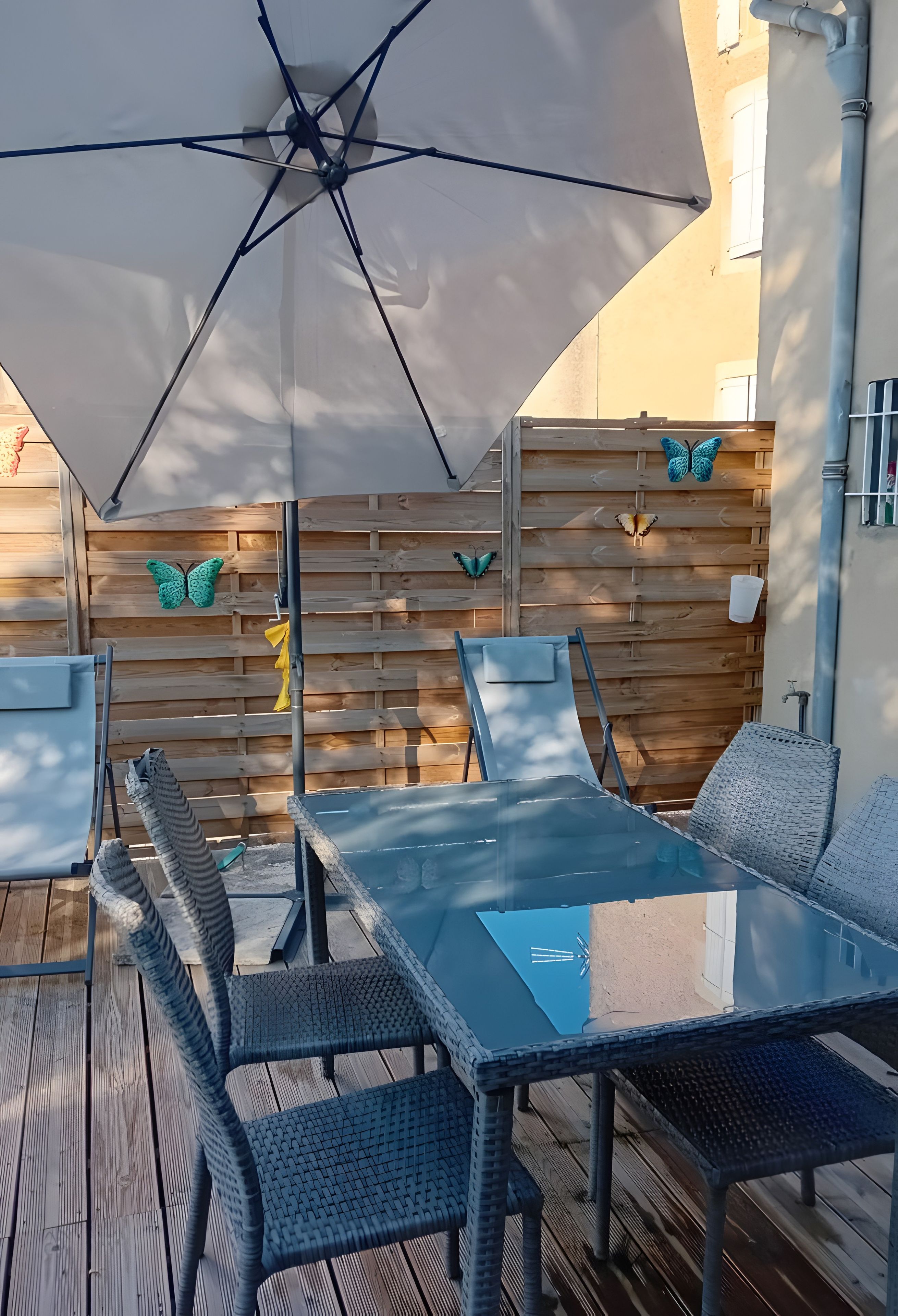 Eine Terrasse im Freien mit einem Glastisch, Stühlen, einem großen Sonnenschirm und dekorativer Schmetterlingswandkunst.
