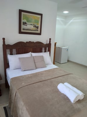 Cama bem arrumada com cabeceira marrom e roupa de cama branca, um radiador branco pequeno e um quadro pendurado na parede.