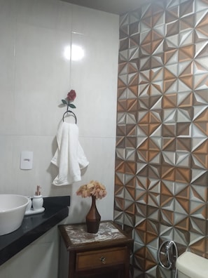Casa de banho com lavatório branco, um porta-toalhas com toalha branca, um vaso com flores, uma mesinha de cabeceira de madeira e uma sanita com chuveiro.