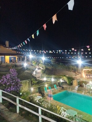 Vista noturna de área de piscina com escadaria que leva a um edifício, cercada por vegetação e uma fileira de bandeiras coloridas.