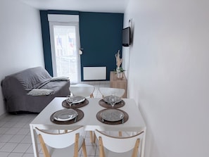 Een moderne woonkamer met een eethoek, een grijze bank, een aan de muur bevestigde televisie en een raam met uitzicht.