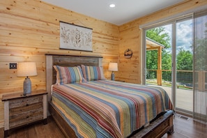 Quarto de cabana de madeira com uma cama, duas luminárias de cabeceira, uma impressão botânica emoldurada e vista do exterior através de uma porta de correr de vidro.