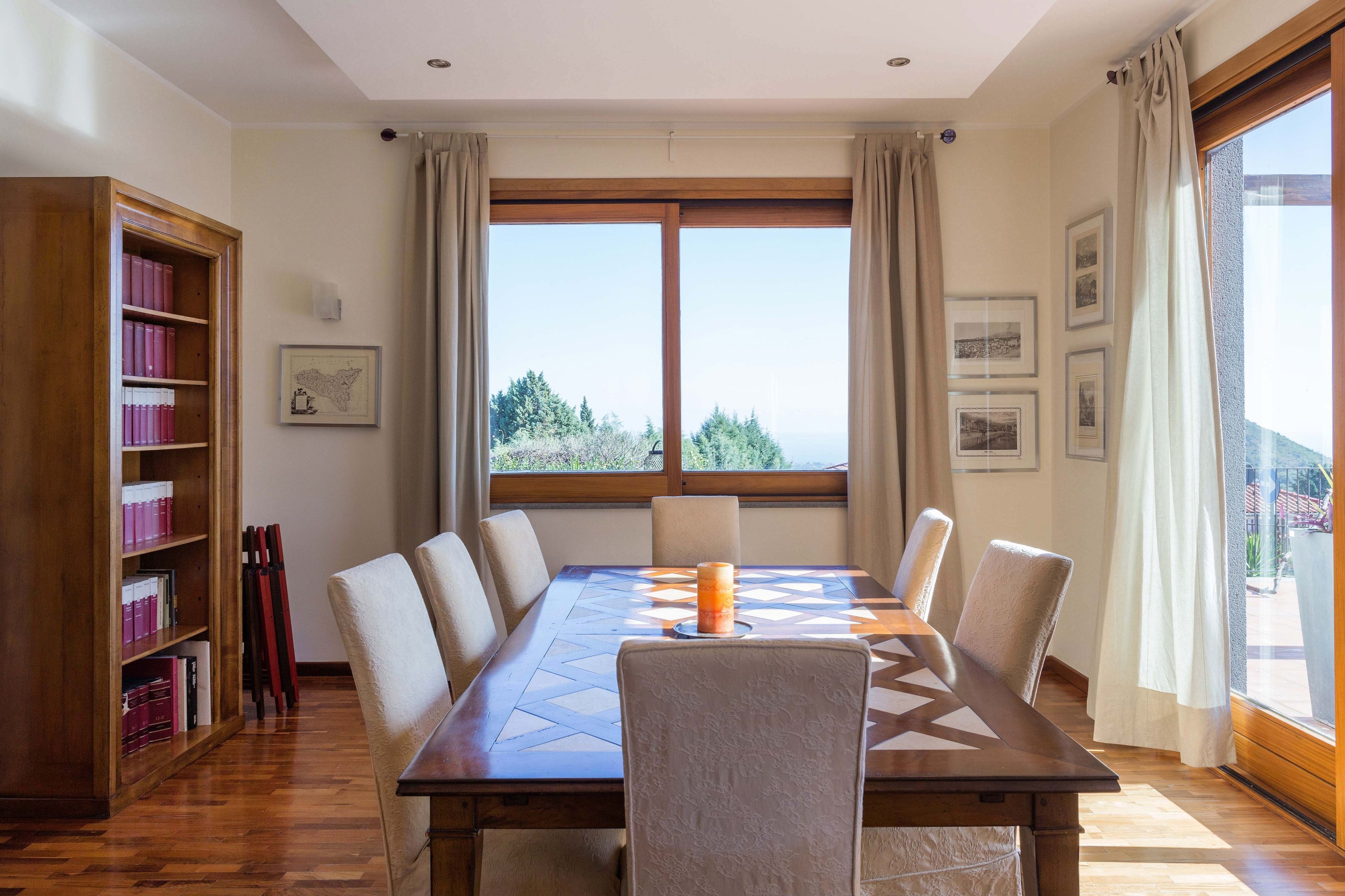 A dining room with a long table, chairs, a bookshelf, and a window with a view.