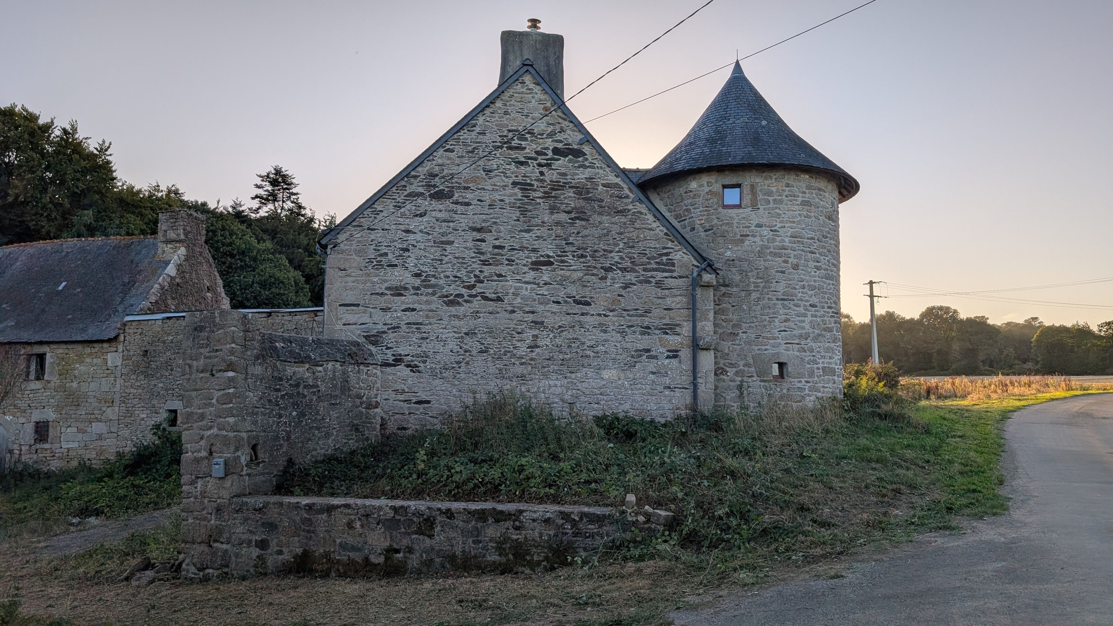 A stone building with a conical roof and a cylindrical tower, surrounded by greenery and a rural road.
