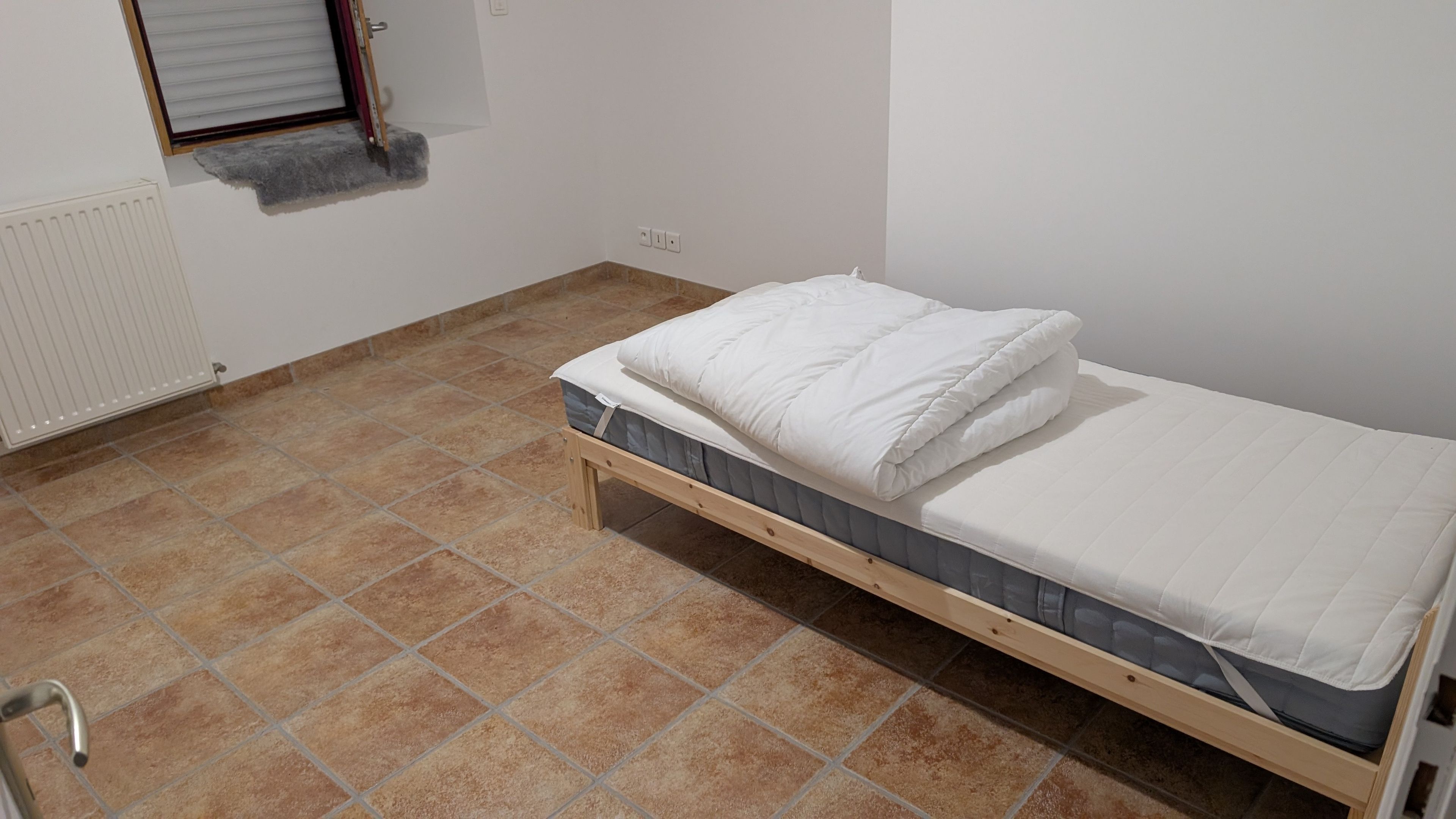 Una habitación pequeña con una cama individual, estructura de madera, colchón gris y ropa de cama blanca.