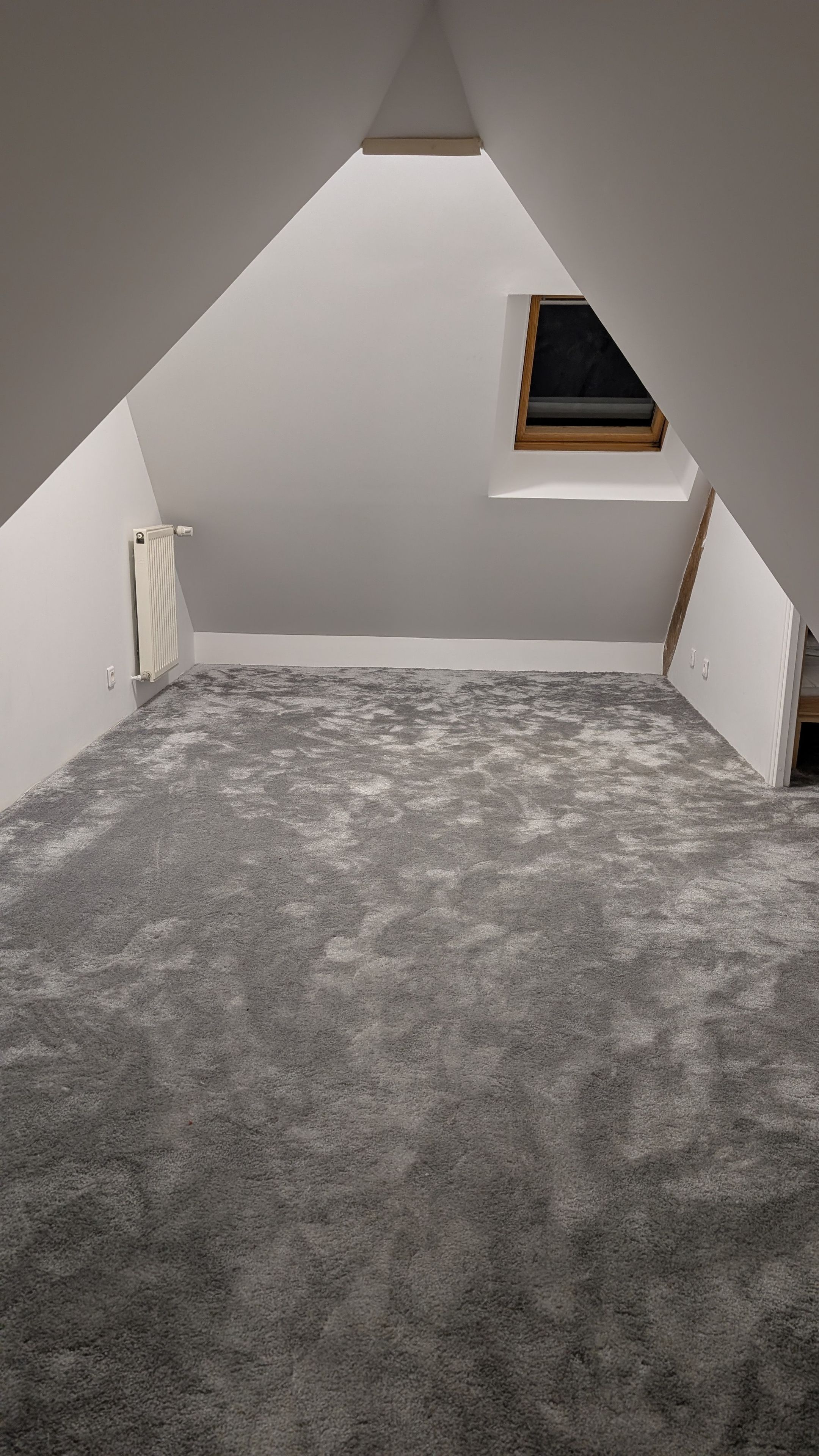 A room with a sloped ceiling, a radiator, a small window, and a grey carpet.