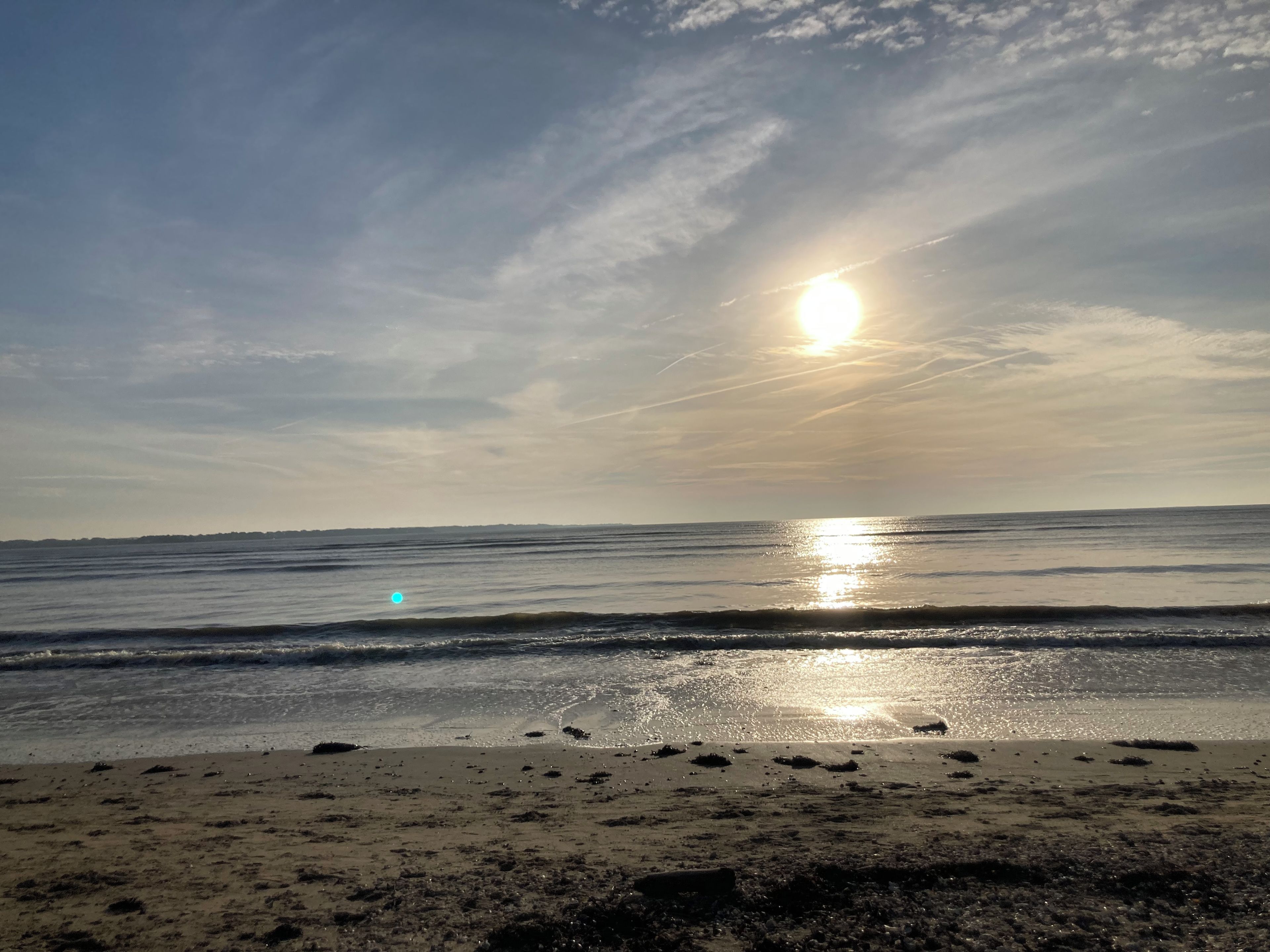 A beach with the sun reflecting on the water, a clear sky, and a distant shoreline.