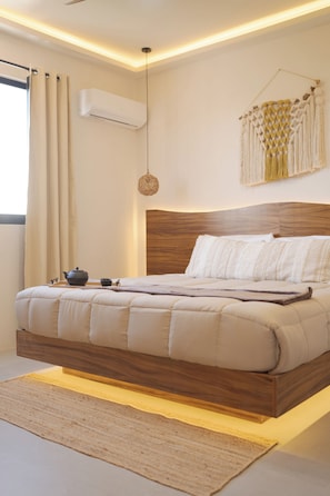 Un dormitorio moderno con cabecera de madera, cama con ropa de cama blanca, decoración de pared de macramé colgante y una mesita de noche con tetera y taza.