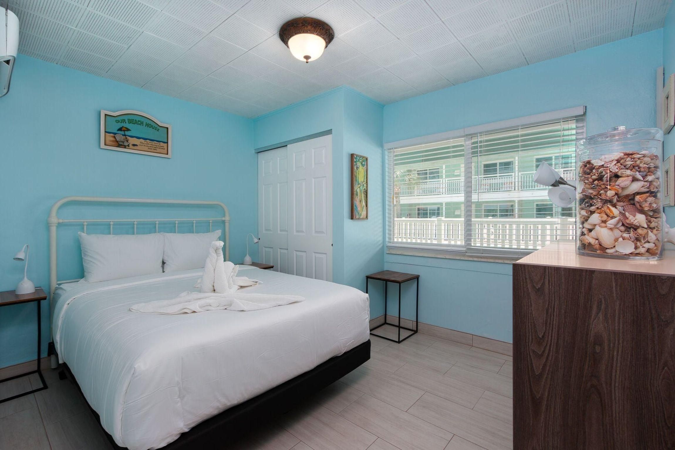 A hotel room with a bed, a bedside table, a nightstand, a window with blinds, and a decorative jar of seashells.