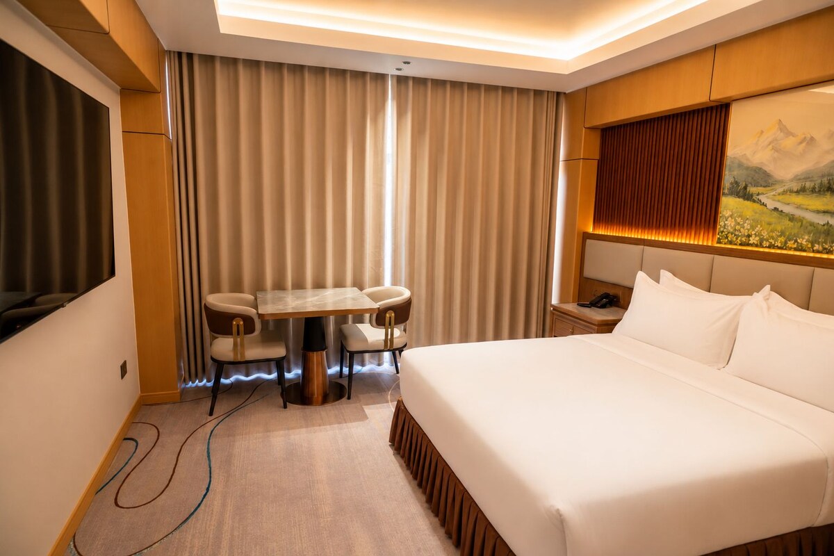 Ein Hotelzimmer mit einem Bett, einem Schreibtisch mit zwei Stühlen, einem Gemälde an der Wand und einem Vorhang.