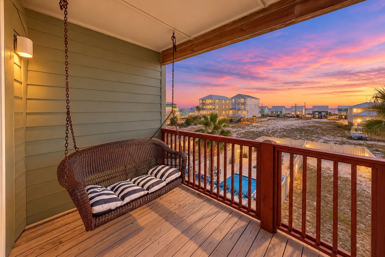 Une terrasse en bois avec un fauteuil suspendu en osier, une piscine et une vue sur les maisons de plage au coucher du soleil.