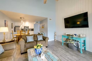 Een woonkamer met een zithoek, een salontafel met bloemen, een televisie en een keuken op de achtergrond.