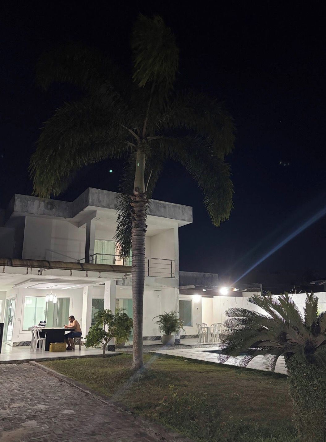 A modern two-story building with a balcony, a palm tree in the foreground, and a person sitting on a bench.