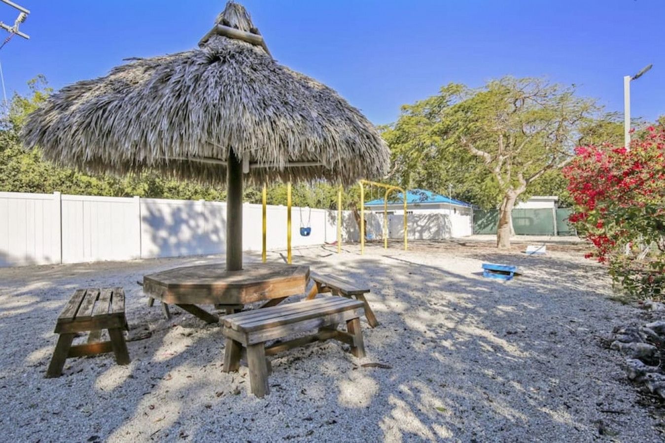 A sandy outdoor area with a thatched umbrella, picnic tables, and a playground structure.