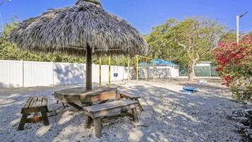 A sandy outdoor area with a thatched umbrella, picnic tables, and a playground structure.
