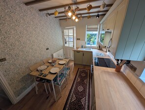 A dining area with a wooden table set for two, a patterned wallpaper, and a kitchen with light-colored cabinets and a window. - Charming Yorkshire Cottage | Log Burner | Dog Friendly | Near Malton (Brawby)