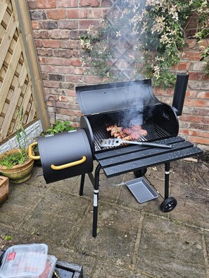 A barbecue grill with meat being cooked, a portable gas tank, and a metal tray on a concrete surface. - Charming Yorkshire Cottage | Log Burner | Dog Friendly | Near Malton (Brawby)
