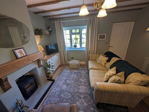 A cozy living room with a fireplace, a sofa, a television, and a window with curtains. - Charming Yorkshire Cottage | Log Burner | Dog Friendly | Near Malton (Brawby)