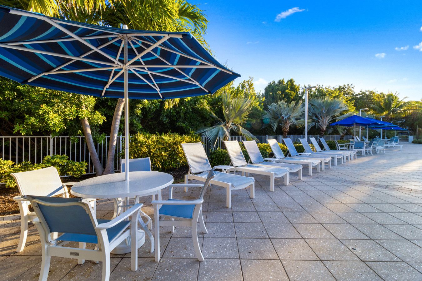 Un espace extérieur lumineux, aménagé avec des chaises longues et des tables blanches, entouré de verdure et de palmiers.